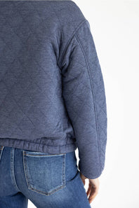 Close-up back view of a person wearing a quilted navy blue jacket with elasticated hem, paired with blue jeans, against a plain white background.