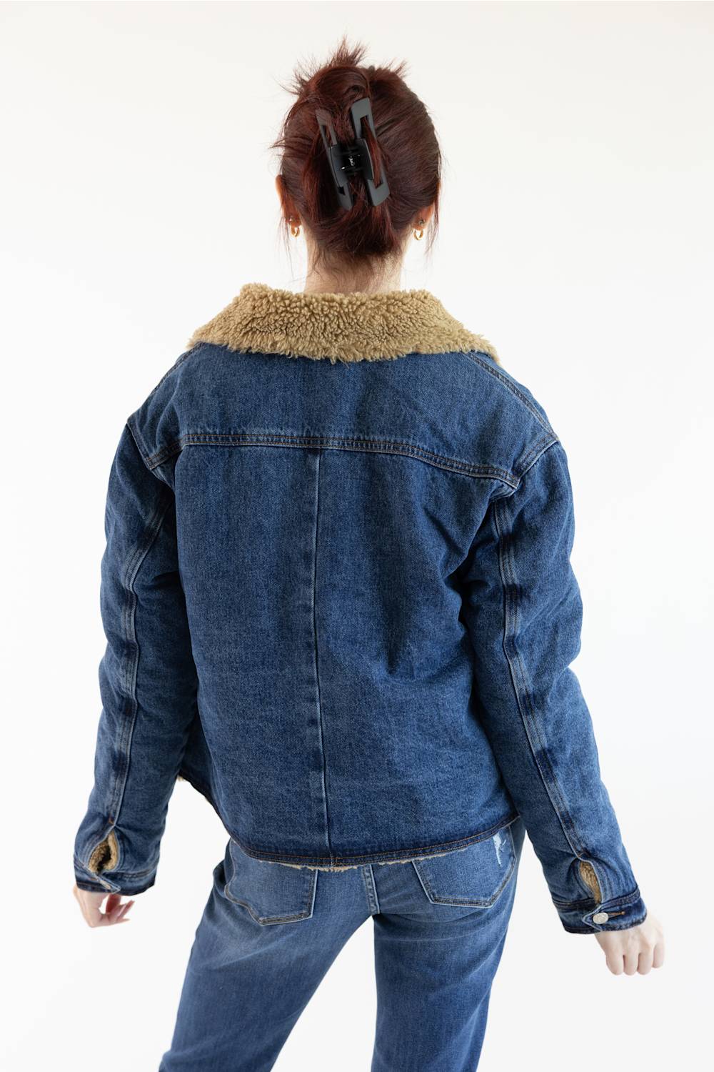A person shown from the back wearing a blue denim jacket with a tan sherpa collar, paired with blue jeans, against a plain white background.