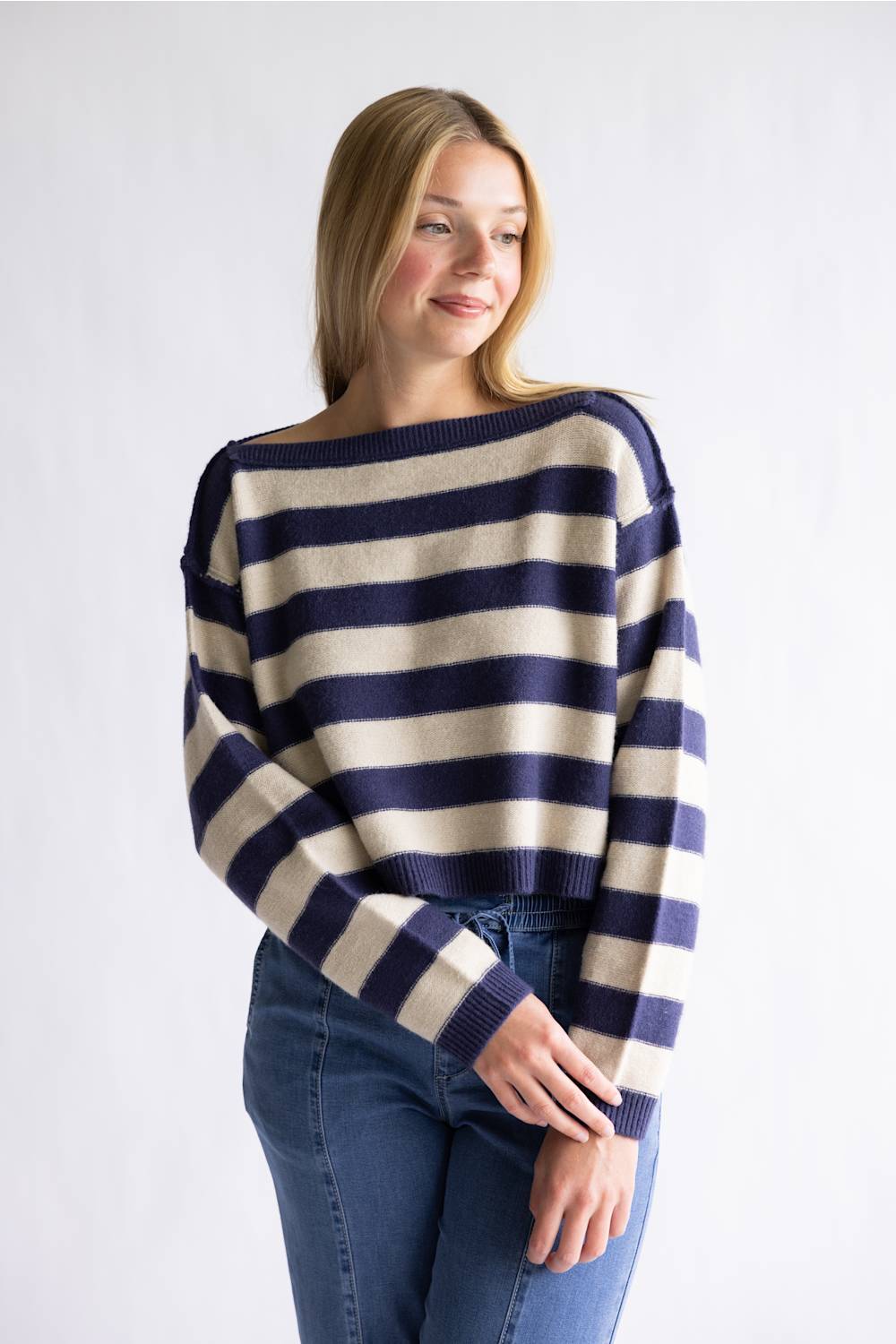 A woman wearing a navy blue and cream striped knit sweater with blue jeans, standing against a plain light gray studio background.