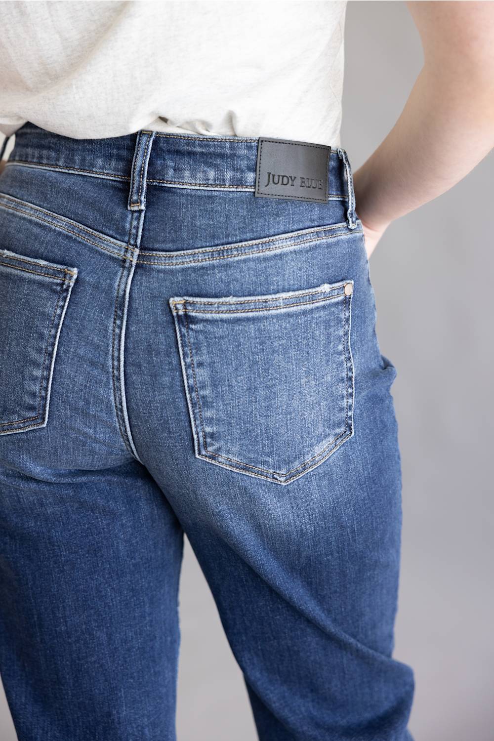 Close-up back view of a person wearing blue denim jeans with a visible Judy Bell brand patch at the waistband, paired with a light heather gray top against a plain light gray background.