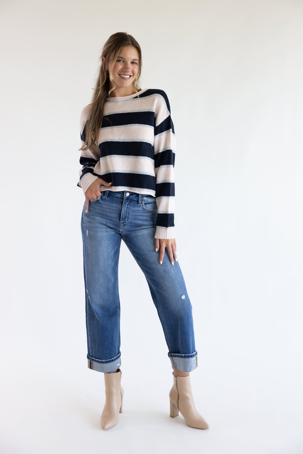 Full-length view of a smiling model wearing the Just Polly long-sleeve knit sweater featuring wide pink and navy horizontal stripes with thin heather grey accents, styled with cuffed straight-leg jeans and beige heel booties against a plain white background.