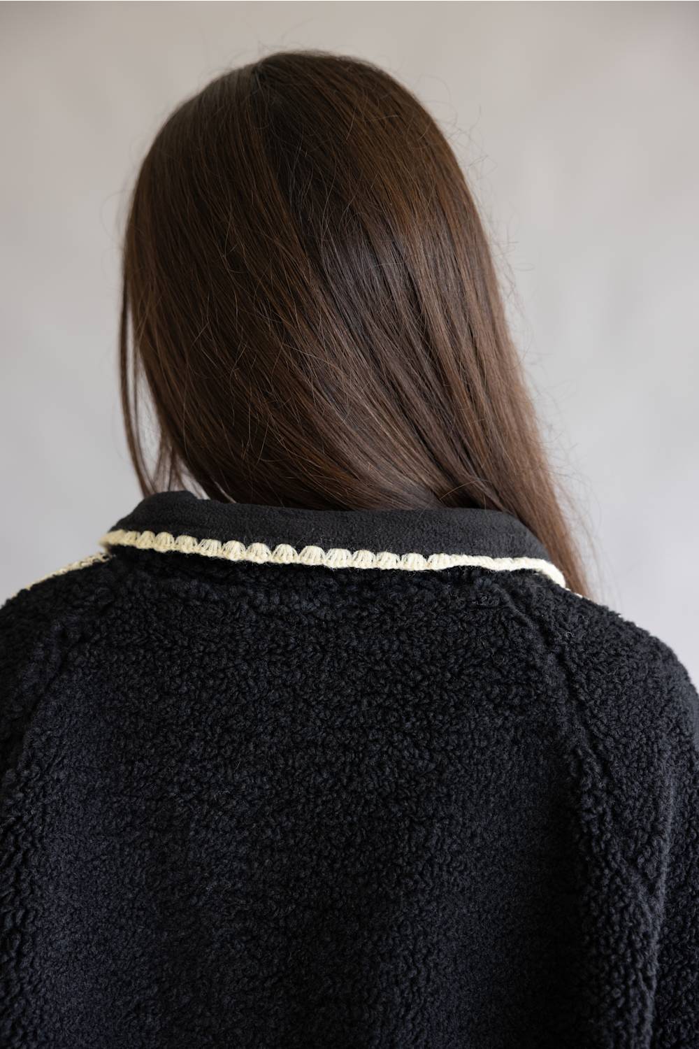 Rear view of a person with long dark brown hair wearing a black textured fleece jacket with a decorative cream beaded collar trim, against a plain light gray background.