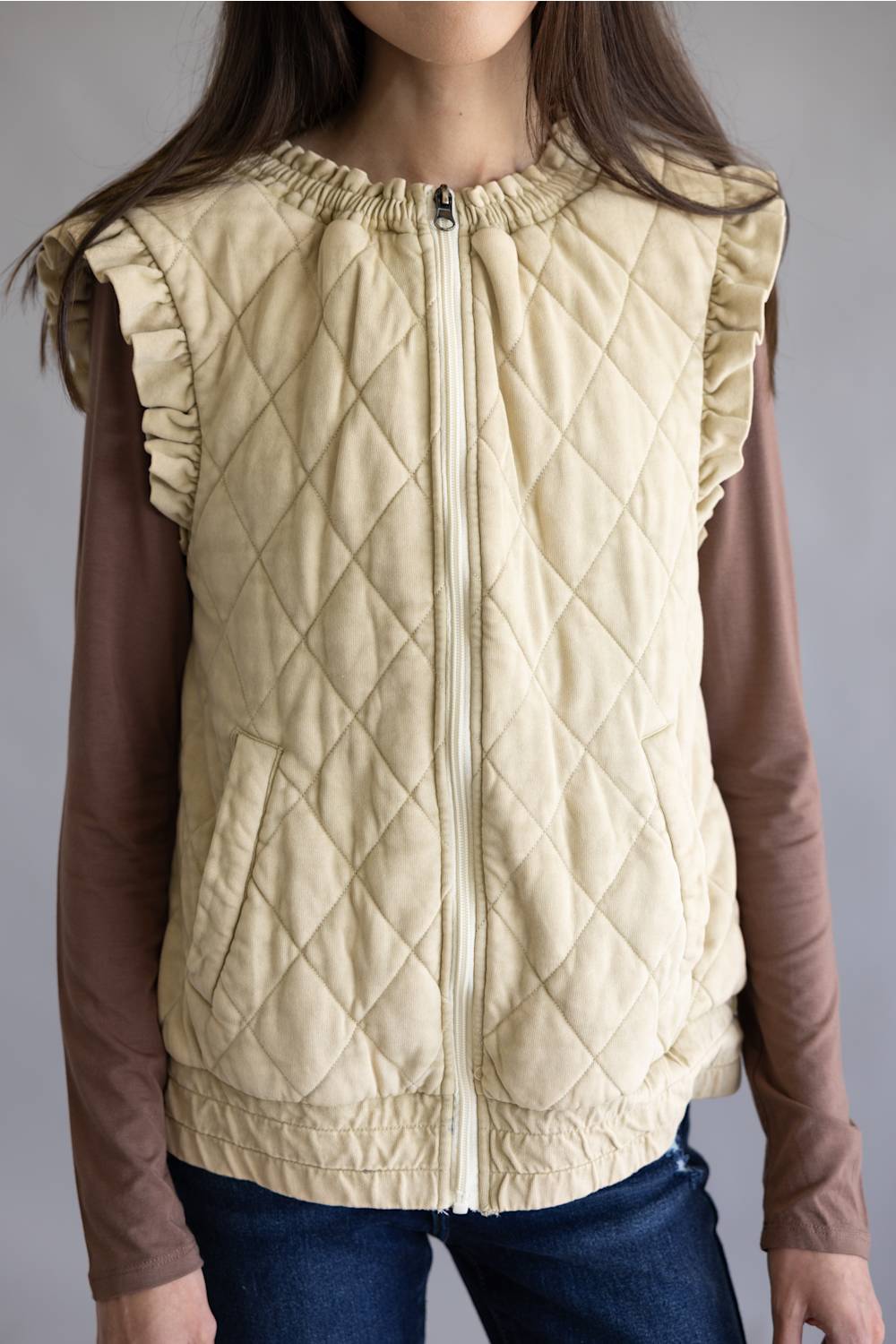 A child wearing a cream-colored diamond-quilted sleeveless vest with ruffled shoulders over a brown long-sleeve shirt and blue jeans, shown from the neck down against a neutral gray background.