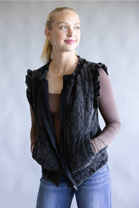 A woman wearing a black quilted sleeveless vest with ruffled shoulders over a brown long-sleeve top and blue jeans, standing against a neutral gray studio background.