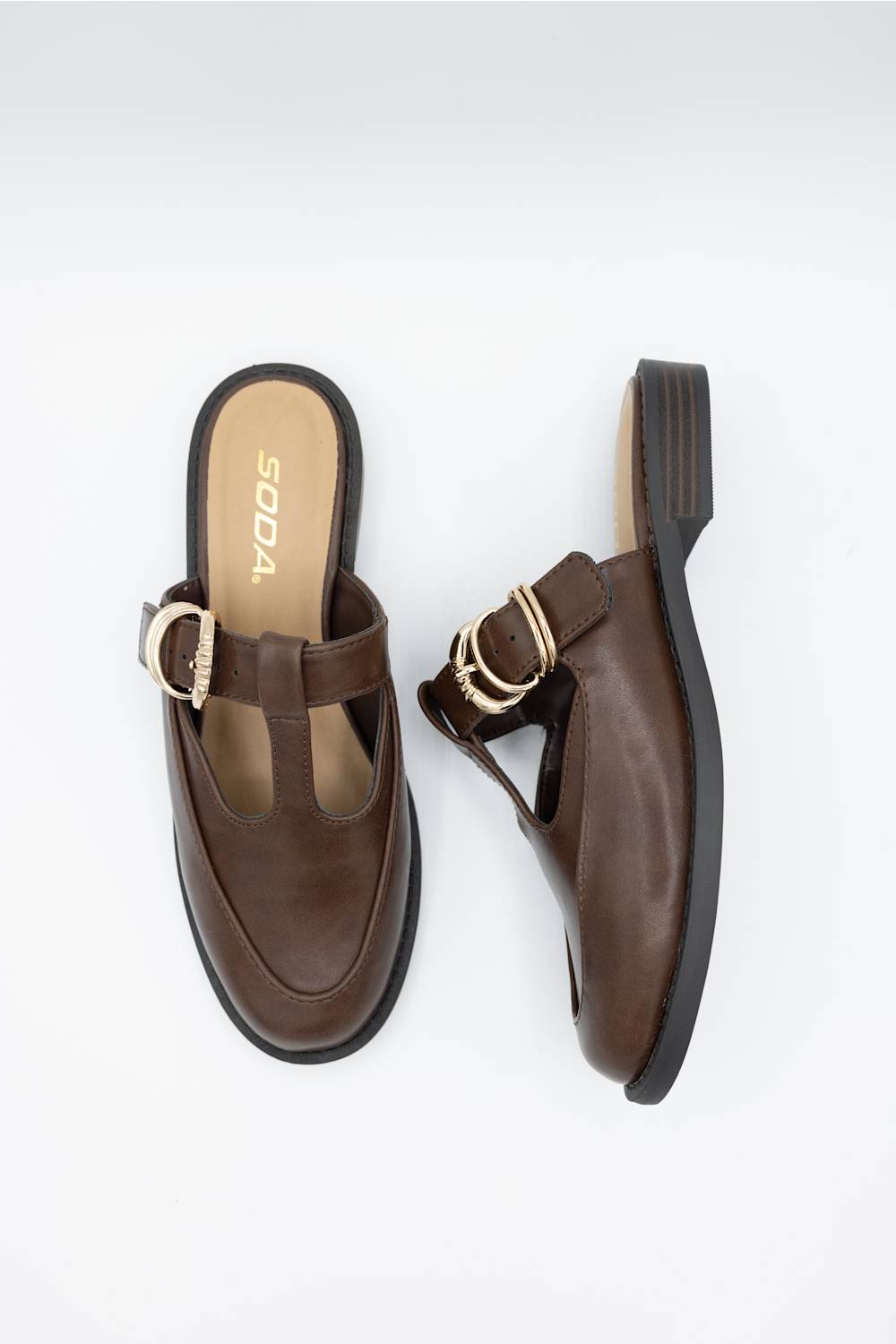 A pair of dark brown leather mule sandals with decorative gold-tone buckle straps, shown from top and side angles on a light gray background. The insole features the brand name 'SODA'.