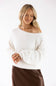 A woman wearing a fuzzy off-the-shoulder white sweater and a brown skirt, standing against a plain white studio background.