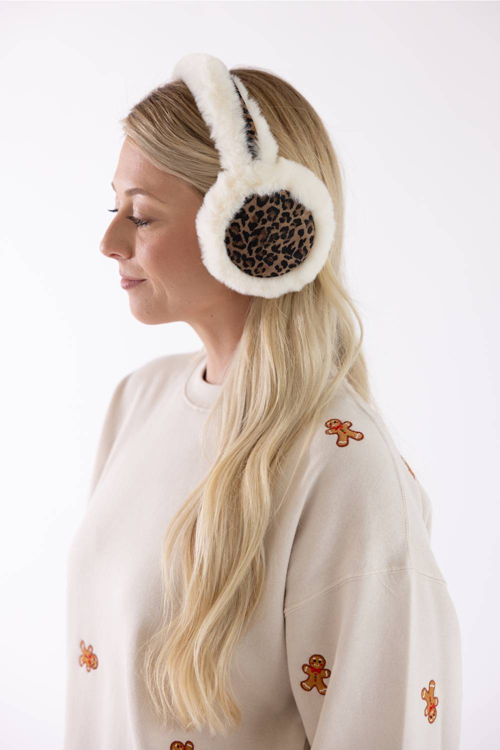 A woman in profile wearing fluffy off-white earmuffs with a brown leopard print center panel, paired with a cream sweatshirt decorated with small gingerbread cookie motifs.