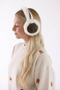 A woman in profile wearing fluffy off-white earmuffs with a brown leopard print center panel, paired with a cream sweatshirt decorated with small gingerbread cookie motifs.