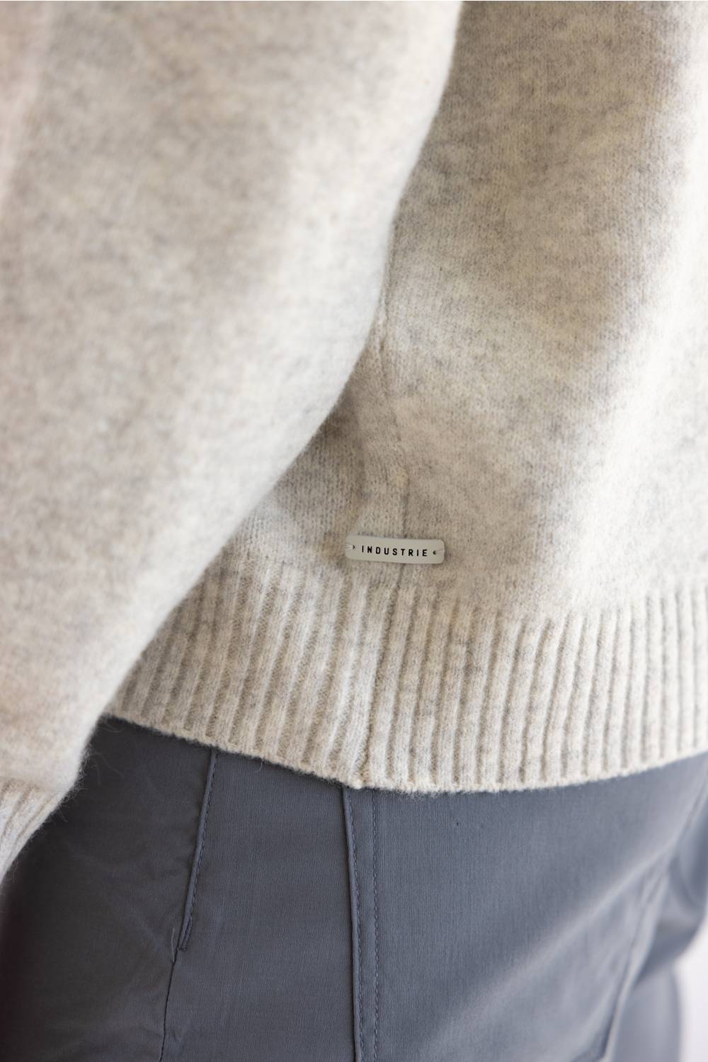 Close-up of a light beige knit sweater with a ribbed hem and a small rectangular brand tag reading INDUSTRIE, worn over dark gray pants.
