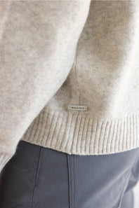Close-up of a light beige knit sweater with a ribbed hem and a small rectangular brand tag reading INDUSTRIE, worn over dark gray pants.