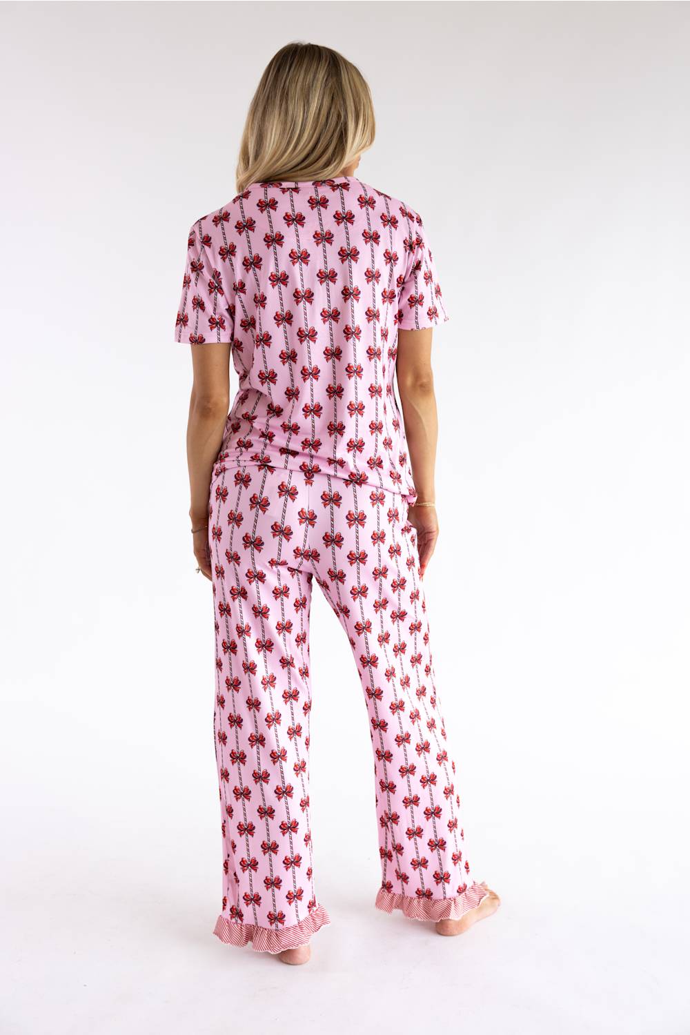A woman viewed from the back wearing a pink short-sleeve pajama top and matching pajama pants with an allover red bow print and ruffled trim at the cuffs, against a plain white studio background.