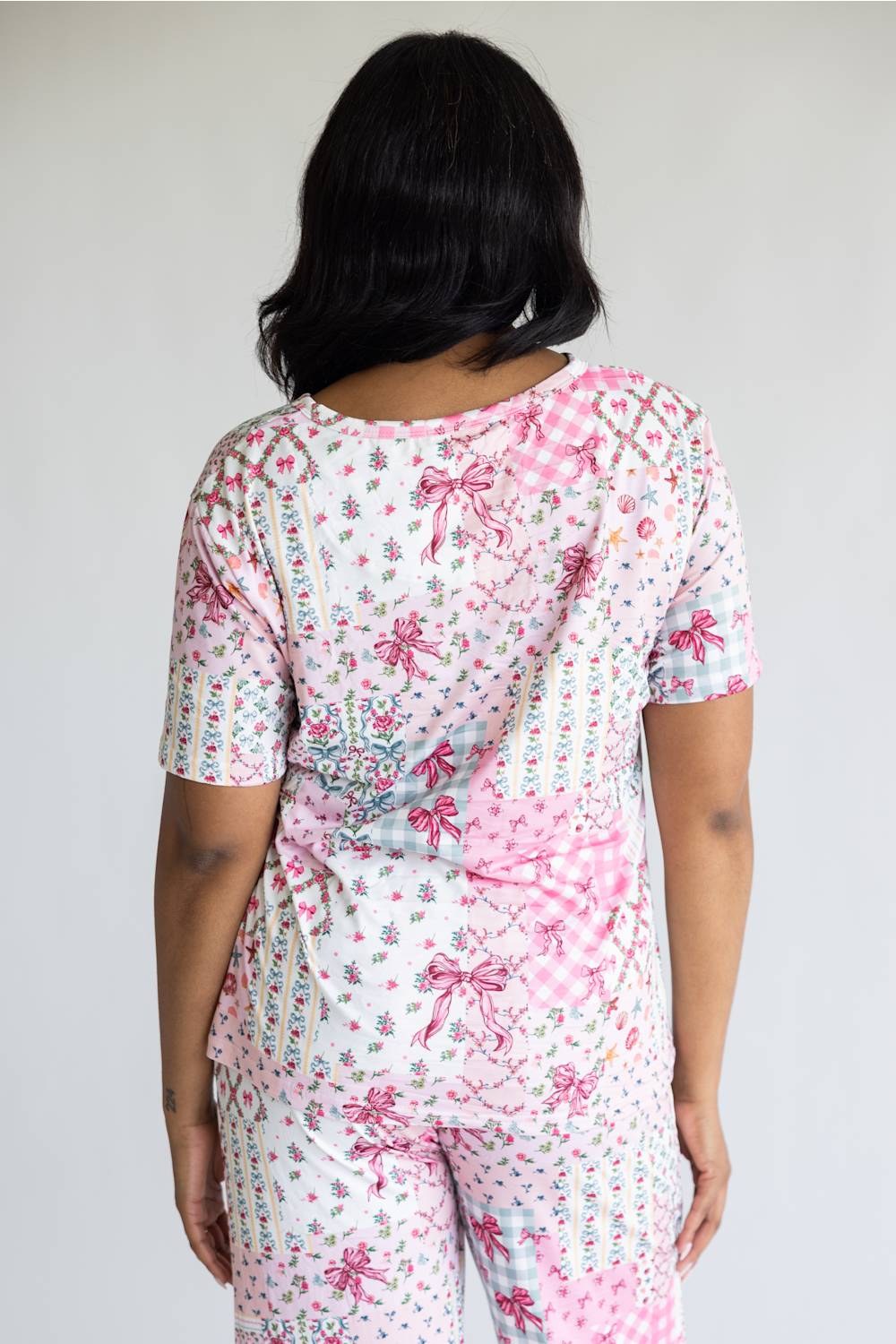 A woman shown from behind wearing a matching short-sleeve pajama set with a pink and white floral print, gingham check, and bow pattern, against a plain light gray background.