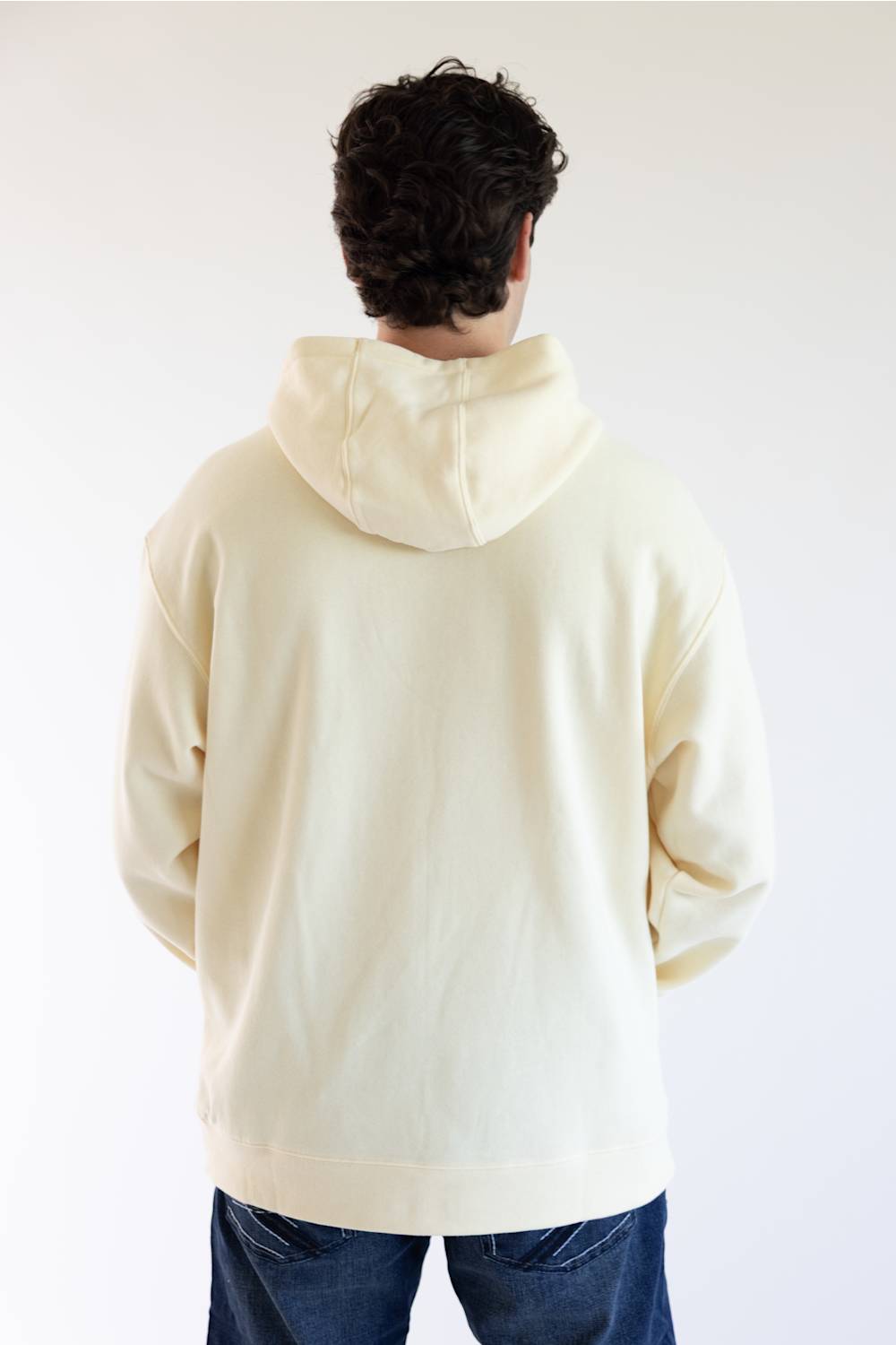 A person viewed from behind wearing an oversized cream-colored hoodie and blue jeans against a plain light background.