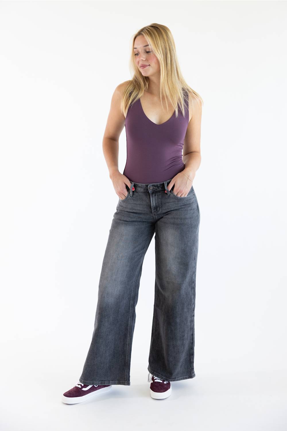 A woman modeling a sleeveless purple tank top and wide-leg gray jeans with burgundy sneakers, standing against a plain white studio background.