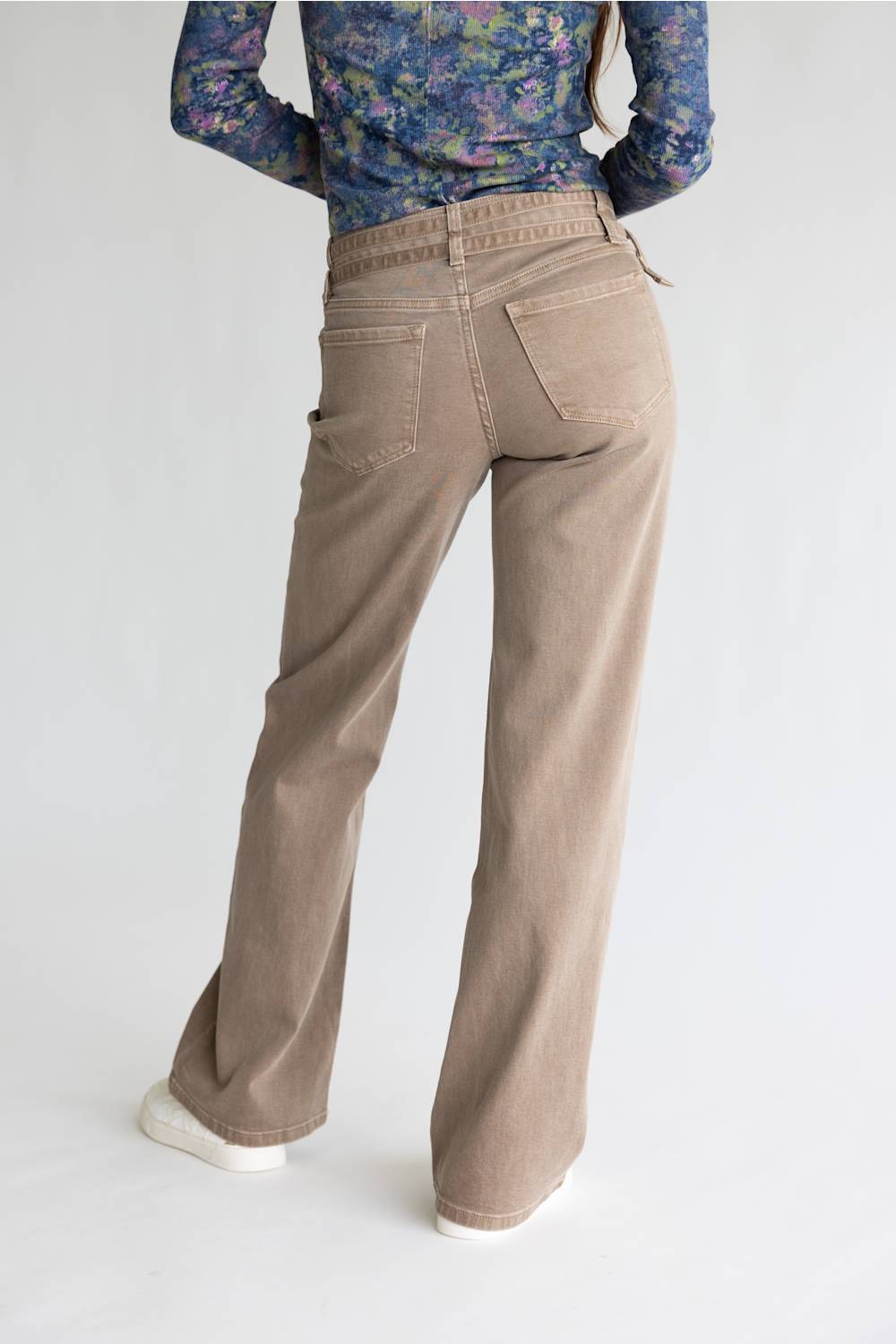 A person shown from the back wearing beige wide-leg jeans, a blue floral long-sleeve top, and white sneakers against a plain light gray studio background.