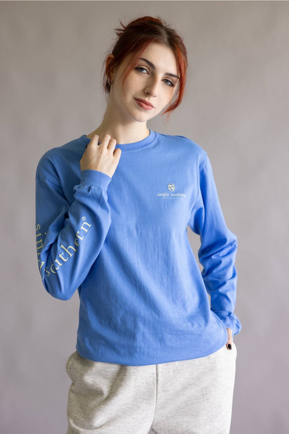 A woman with red hair wearing a blue long-sleeve crewneck sweatshirt with gold text on the sleeve and a small crest logo on the chest, paired with light gray sweatpants, against a neutral gray background.