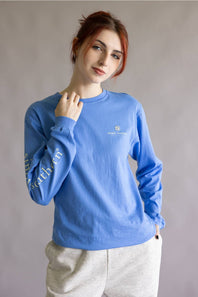 A woman with red hair wearing a blue long-sleeve crewneck sweatshirt with gold text on the sleeve and a small crest logo on the chest, paired with light gray sweatpants, against a neutral gray background.