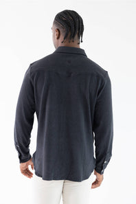 A man viewed from behind wearing a dark charcoal gray long-sleeve button-up shirt and white pants, standing against a plain white studio background.