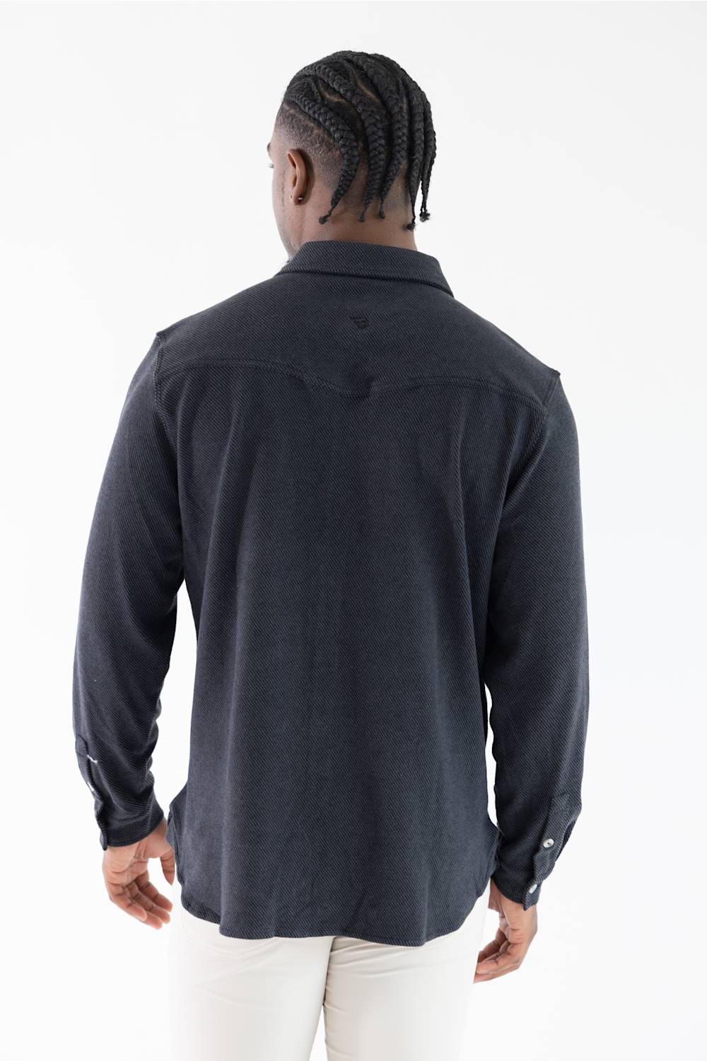 A man viewed from behind wearing a dark charcoal gray long-sleeve button-up shirt and white pants, standing against a plain white studio background.
