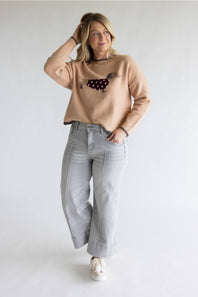 A woman modeling a beige crewneck sweater with a dachshund graphic, paired with light gray wide-leg jeans and white sneakers, against a plain white studio background.