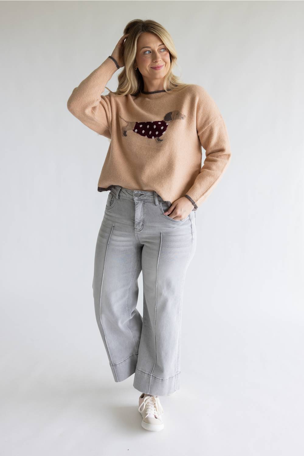 A woman modeling a beige crewneck sweater with a dachshund graphic, paired with light gray wide-leg jeans and white sneakers, against a plain white studio background.