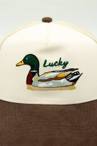 An off-white and brown trucker-style cap featuring an embroidered mallard duck and the word 'Lucky' on the front panel.