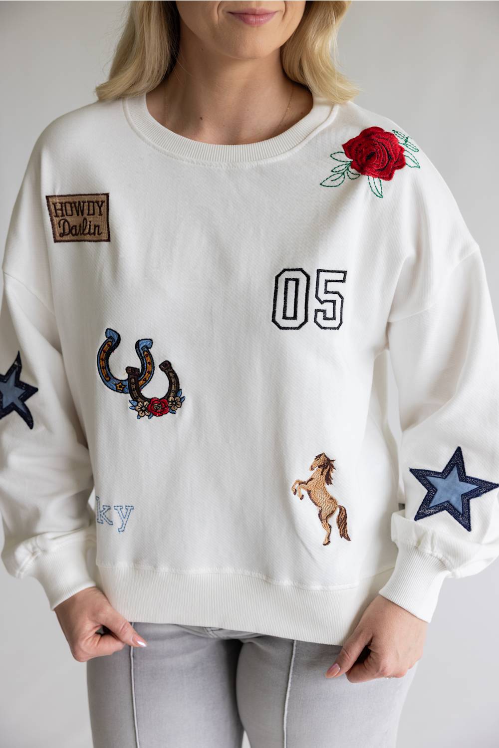 A woman wearing a white crewneck sweatshirt decorated with embroidered patches, including a red rose, a horseshoe, a horse, a star, and the number '05'.