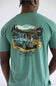 A photo taken from the back of a man in a light green shirt with a graphic of a truck with dogs, a deer carcass, and a cooler in the bed.