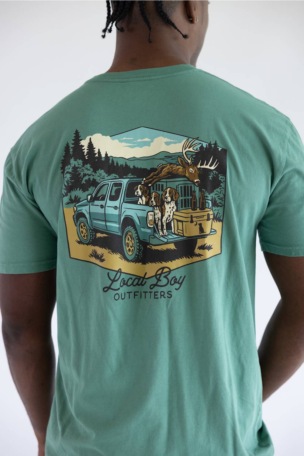 A photo taken from the back of a man in a light green shirt with a graphic of a truck with dogs, a deer carcass, and a cooler in the bed.