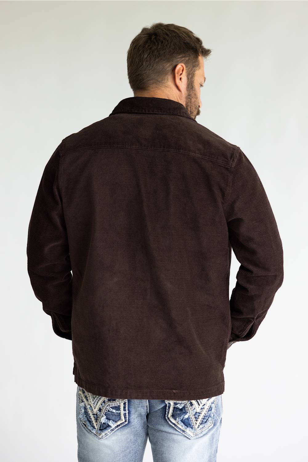 A man viewed from behind wearing a dark brown corduroy jacket and light blue jeans with western-style embroidery on the back pockets, set against a plain light gray background.