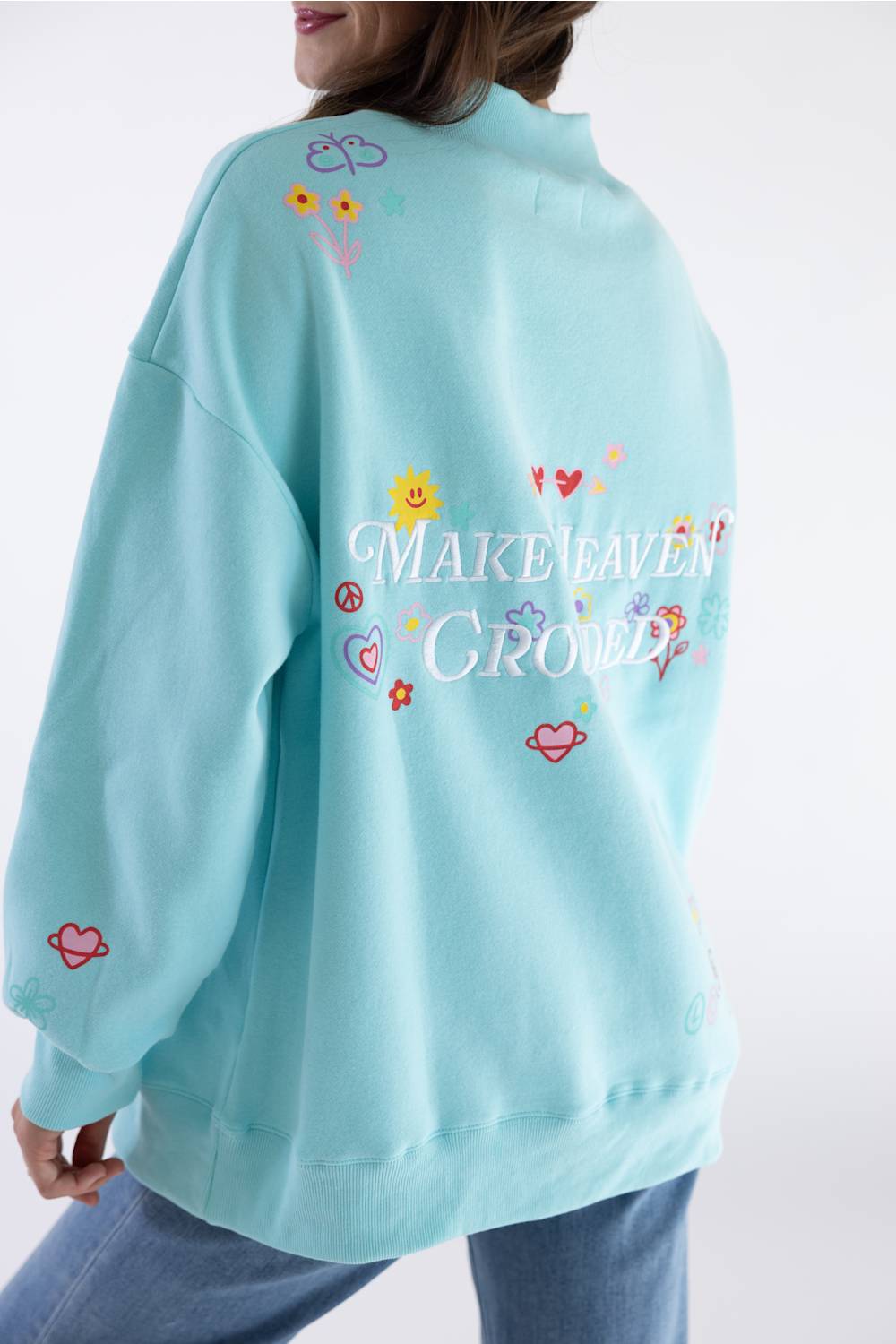 A person wearing an oversized light blue sweatshirt, shown from the back. The sweatshirt features colorful embroidered flowers, hearts, and the phrase 'MAKE HEAVEN CROWded' in white script, with peace signs and small decorative motifs.