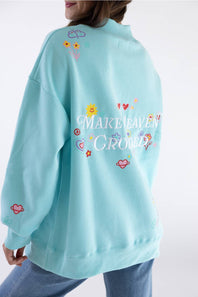 A person wearing an oversized light blue sweatshirt, shown from the back. The sweatshirt features colorful embroidered flowers, hearts, and the phrase 'MAKE HEAVEN CROWded' in white script, with peace signs and small decorative motifs.