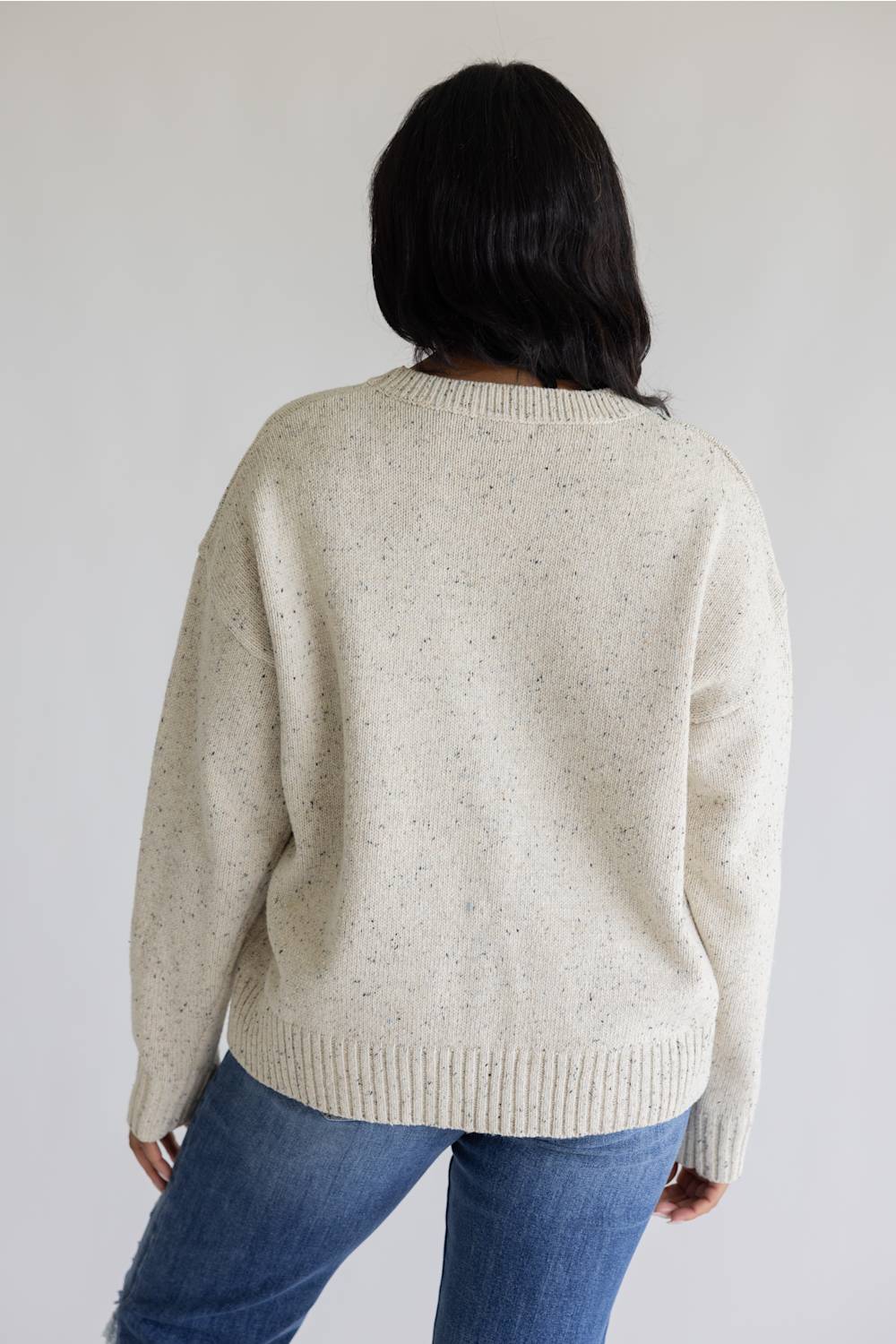 A person shown from the back wearing an oversized cream-colored knit sweater with subtle speckles and blue jeans, set against a plain light gray background.