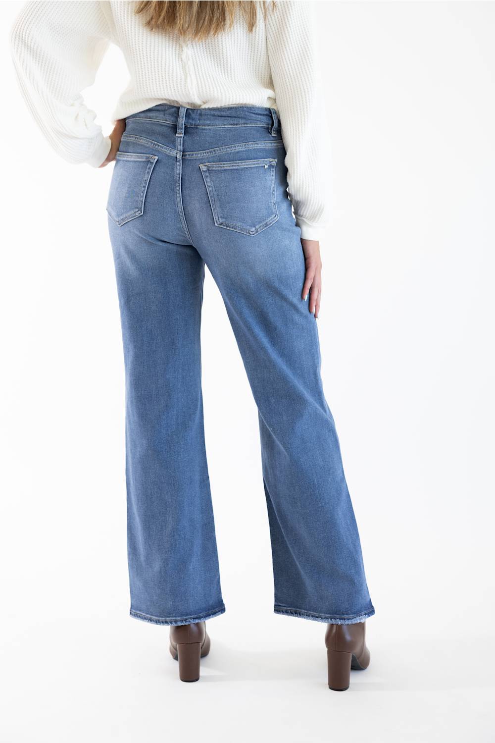 A person shown from the back wearing blue wide-leg jeans, a white ribbed long-sleeve top, and brown heeled shoes against a plain white studio background.
