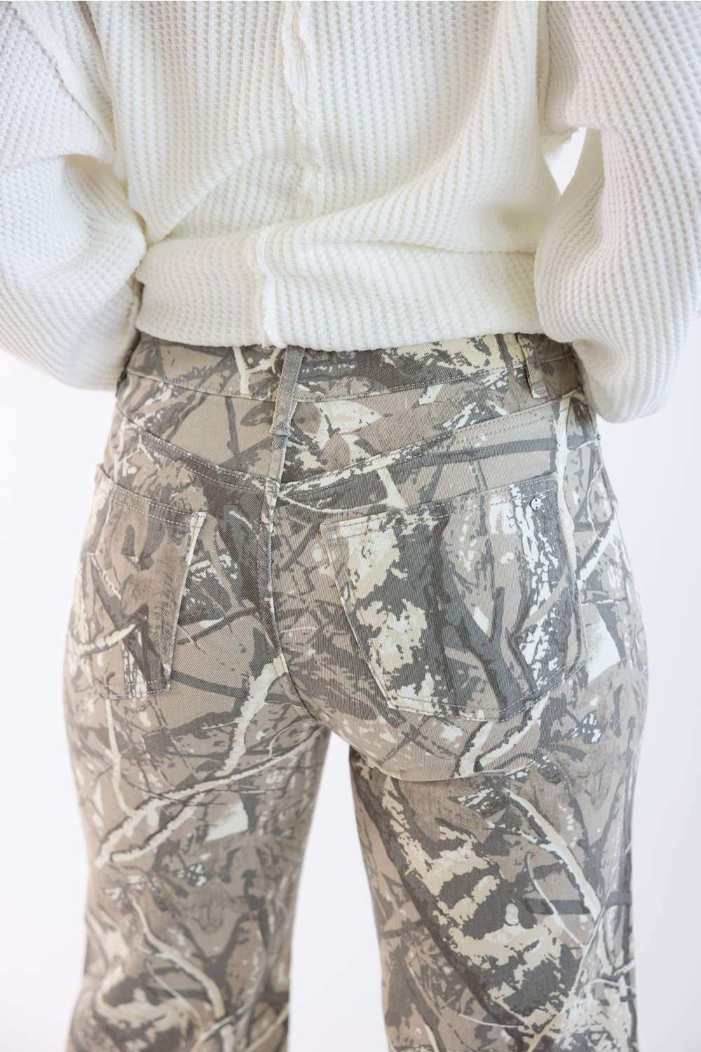 Close-up back view of a person wearing camouflage pants and a cream ribbed sweater against a plain light background.