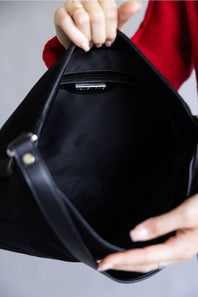 A close-up of a black handbag being held open by hands, showing the interior lining and a small interior label. The person is wearing a red sweater, and the background is a soft light gray.