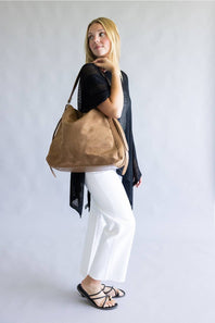 A tan suede shoulder bag with a matching adjustable strap and silver-tone hardware, worn by a model wearing a black mesh top and white pants against a light gray background.
