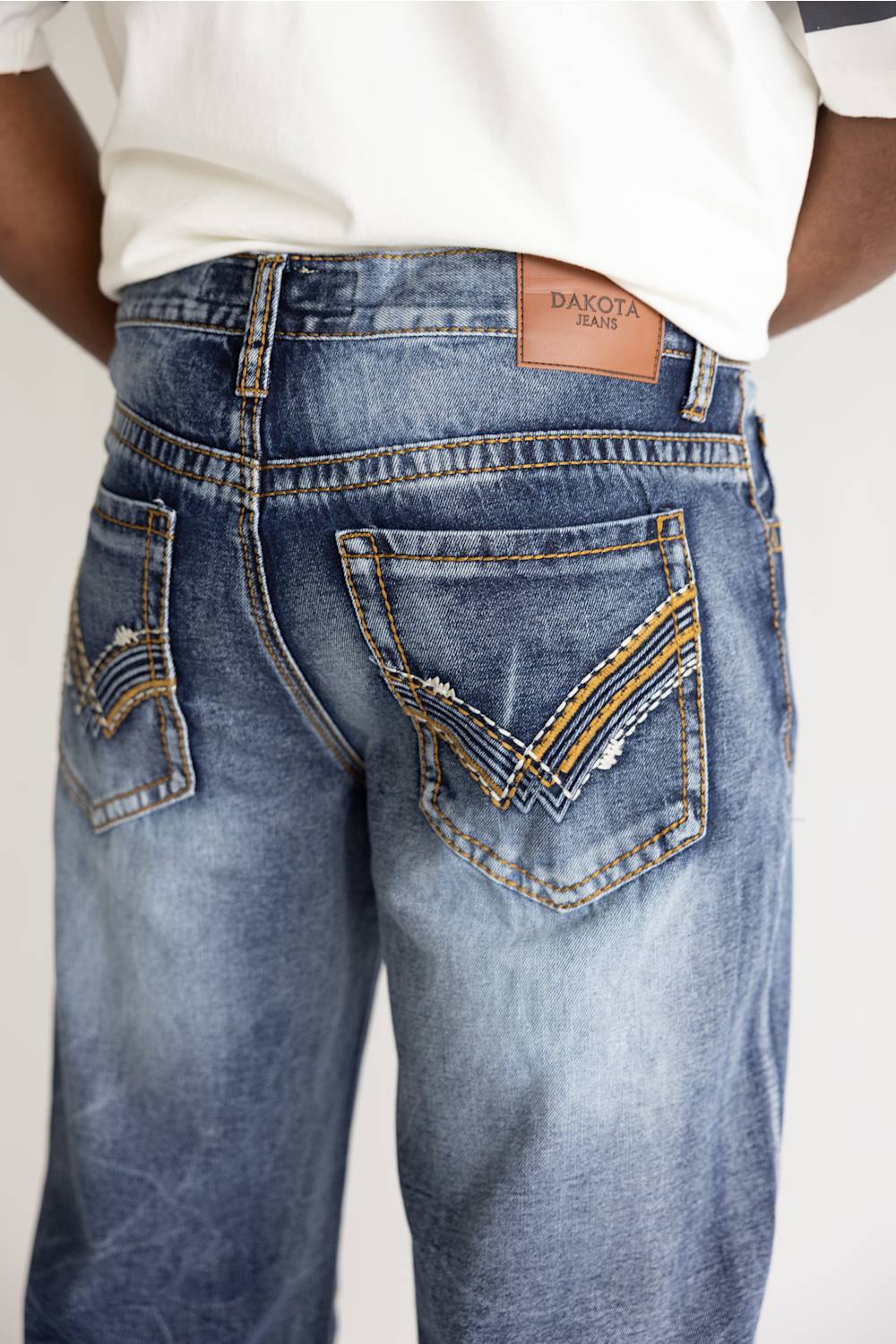Close-up back view of a person wearing blue denim jeans with decorative contrast stitching and a tan leather patch reading DAKOT jeans.
