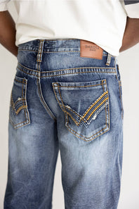 Close-up back view of a person wearing blue denim jeans with decorative contrast stitching and a tan leather patch reading DAKOT jeans.