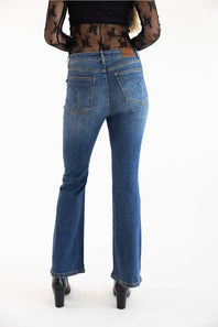 Rear view of a model wearing blue high-waisted flare jeans with a brown leather patch on the back waistband, paired with a black sheer lace top and black heels, against a white studio background.