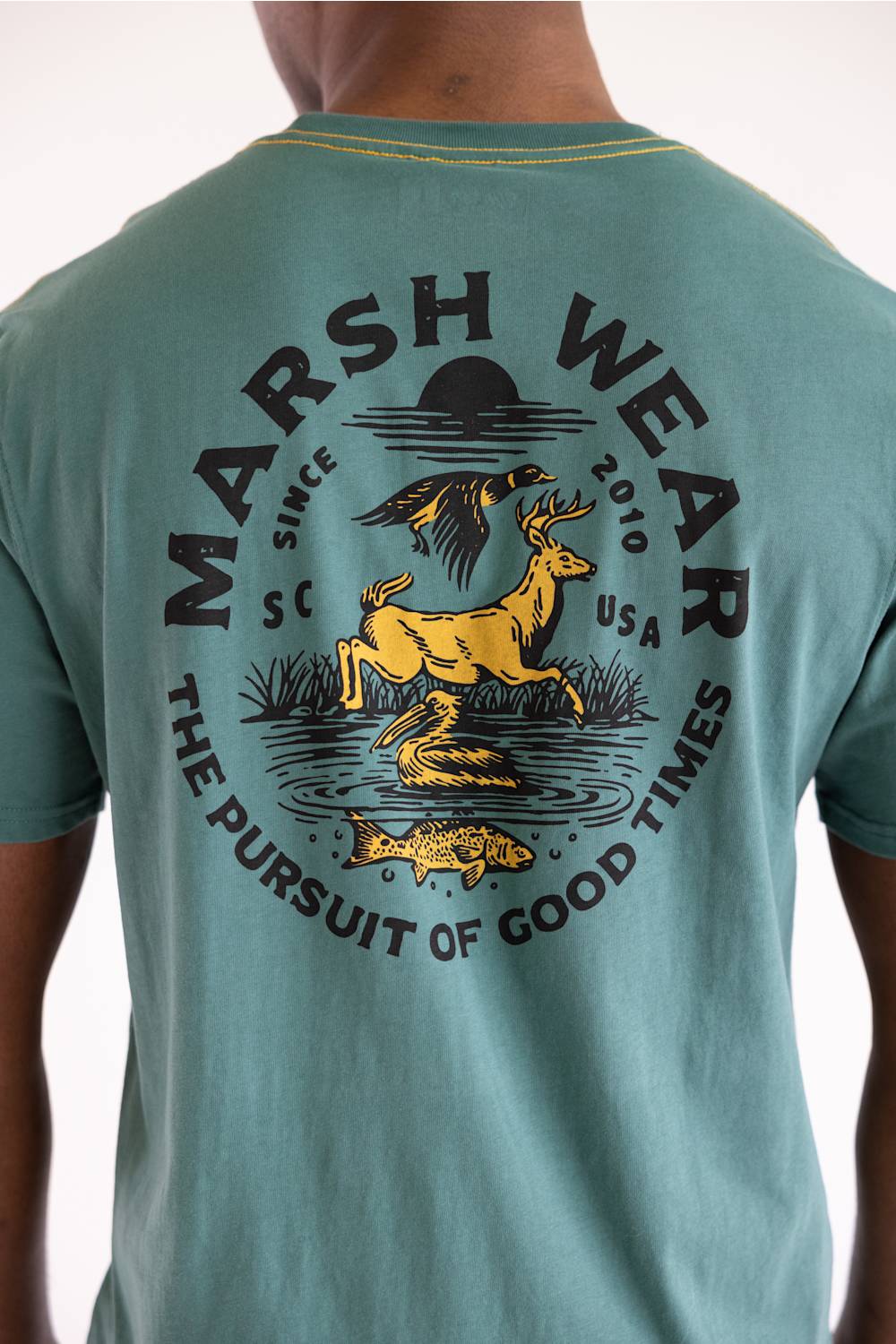 A person wearing a teal short-sleeve t-shirt, viewed from the back. The shirt features a large graphic print in black and yellow on the back, depicting a deer, duck, fish, and the text 'MARSH WEAR' and 'THE PURSUIT OF GOOD TIMES'.