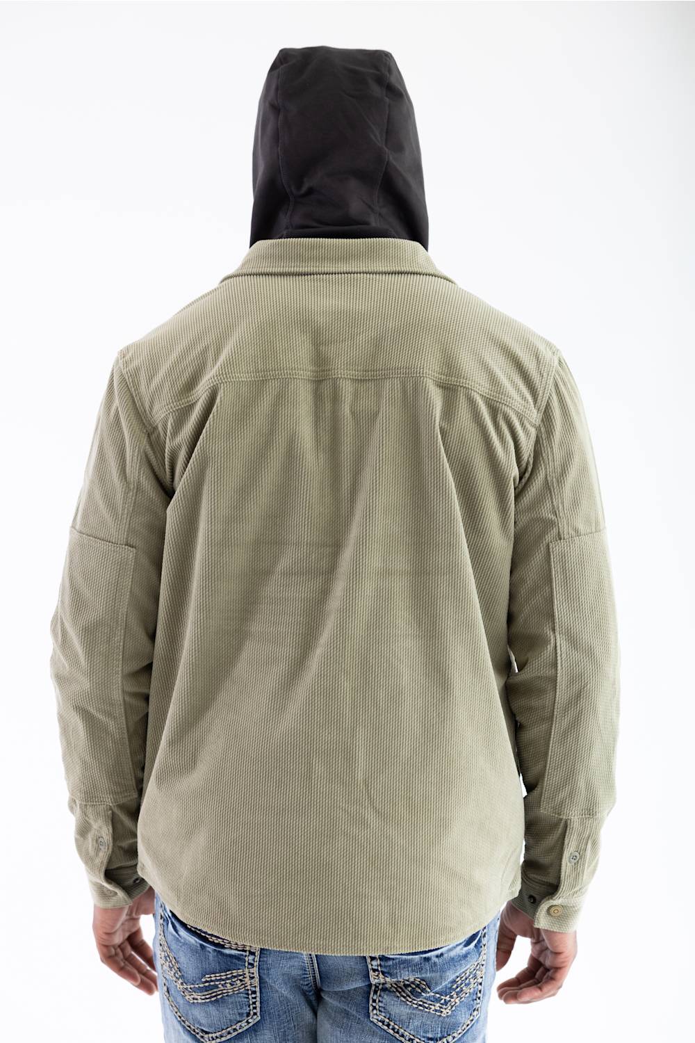 Back view of a model wearing the Marsh Wear Delano Cord Shacket in Sage Brush green corduroy, showing the back yoke and cuff button details, worn over a dark hooded sweatshirt with the hood up, paired with light-wash blue jeans.