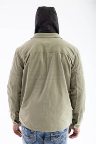 Back view of a model wearing the Marsh Wear Delano Cord Shacket in Sage Brush green corduroy, showing the back yoke and cuff button details, worn over a dark hooded sweatshirt with the hood up, paired with light-wash blue jeans.