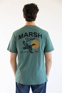 A man viewed from behind wearing a teal green t-shirt with a large black graphic on the back reading 'MARSH Wear' and 'EST. 2010 USA', featuring a flying duck and a circular gold emblem.