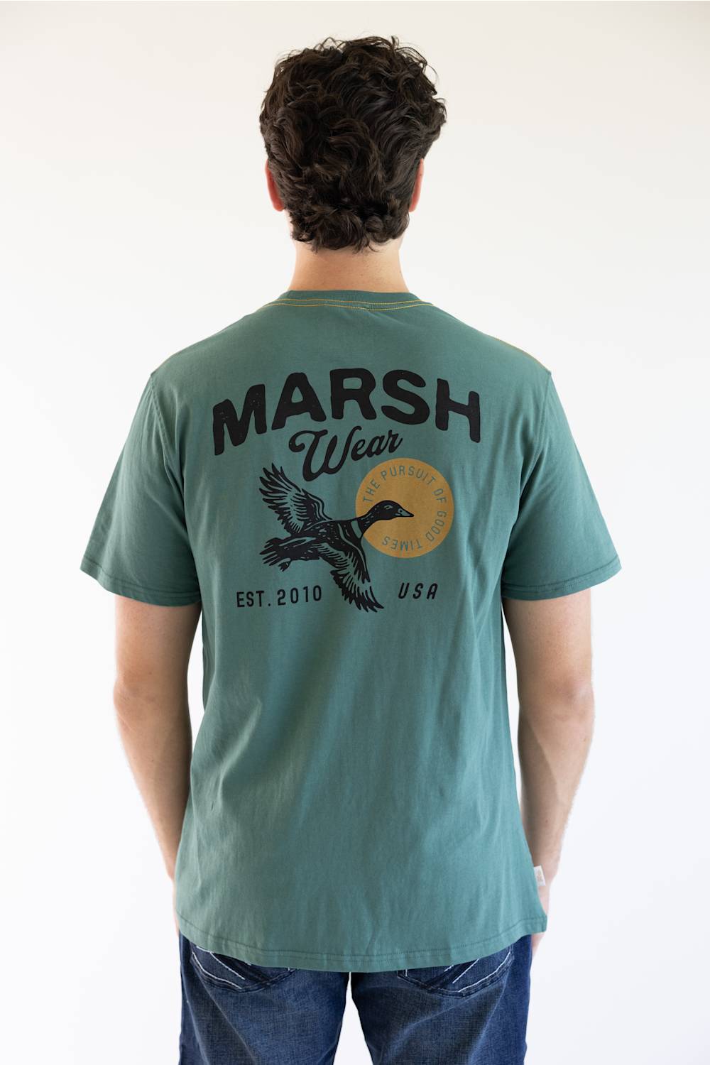 A man viewed from behind wearing a teal green t-shirt with a large black graphic on the back reading 'MARSH Wear' and 'EST. 2010 USA', featuring a flying duck and a circular gold emblem.