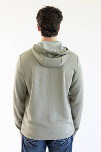 A man wearing a Marshwear hooded quarter-zip pullover in a textured sage green fabric with a small orange zipper and a small Marshwear  logo patch on the chest, paired with blue jeans against a plain white background.