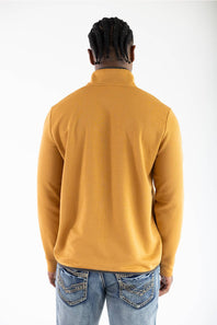 Back view of a model wearing the Buckthorn Brown Marsh Wear Sullivan Quarter Zip Pullover, showing the textured ribbed fabric construction, dark trim along the hem, and stand-up collar, paired with light-wash blue jeans featuring decorative pocket stitching.