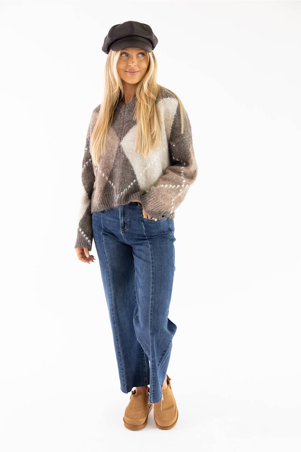 Front view of a smiling woman wearing a brown Miracle argyle V-neck sweater featuring a soft fuzzy knit fabric and an all-over diamond pattern, paired with dark denim jeans, brown ankle boots, and a dark brown baker boy cap.