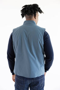 A man viewed from behind wearing a slate blue sleeveless vest over a dark navy long-sleeve shirt and blue jeans, against a plain white studio background.