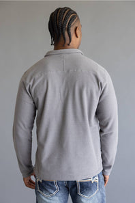 A man viewed from behind wearing a light gray long-sleeve knit polo shirt and blue jeans, standing against a plain gray studio background.